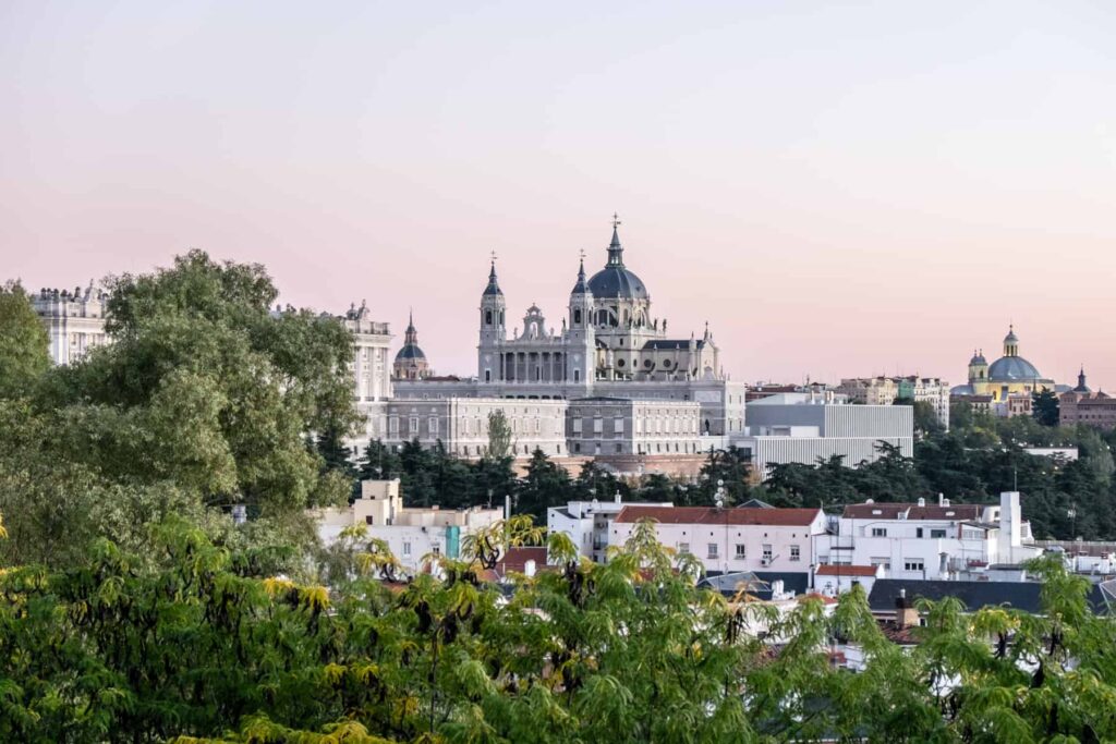A silvery white palace complex with dark grey dome roof and spires is elevated about square city buildings in Madrid. The view is from within the foliage of parkland, and at a distance.